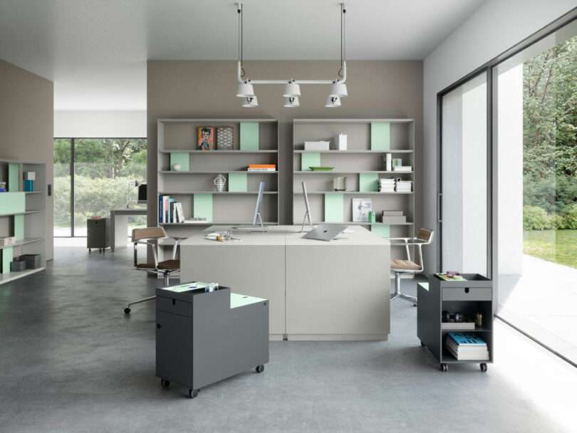 Modern office space with large windows, minimalist desks, open shelving, and rolling storage units on a concrete floor. Natural light fills the room.