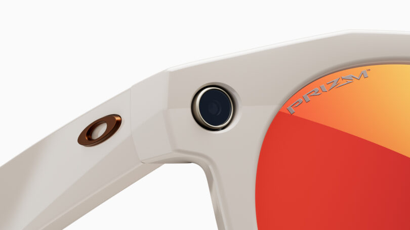 Close-up of the hinge of a white Meta sunglasses frame with an embedded small camera and orange-tinted lens labeled 