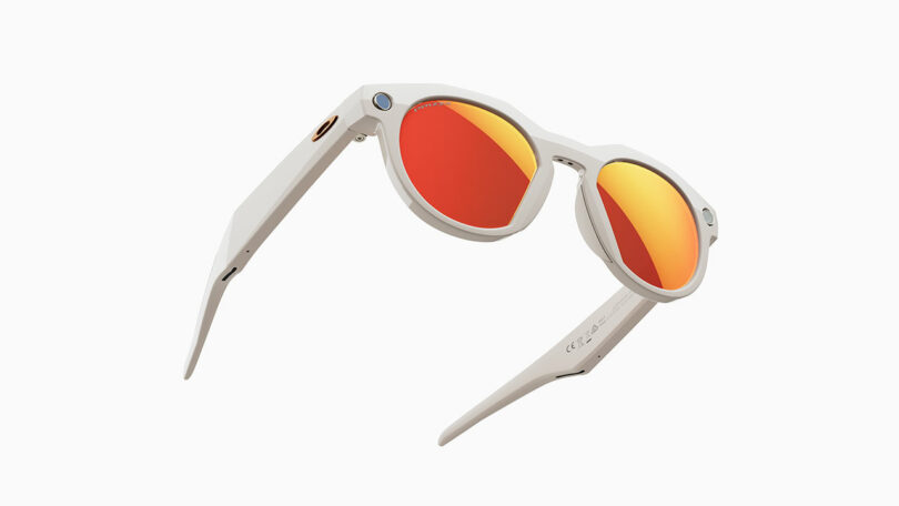 Meta white smart sunglasses with round frames and red-orange mirrored lenses, featuring a small camera and button on the temples, are shown on a white background.