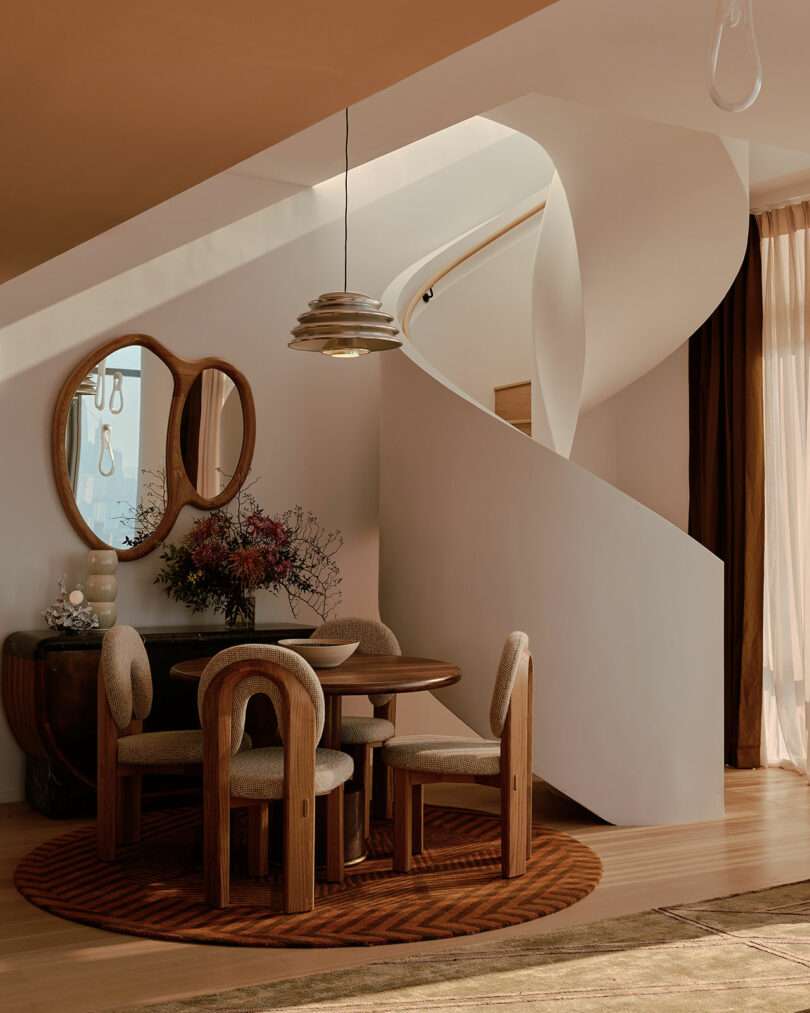 A modern dining area with a round table and chairs on a circular rug. A spiral staircase ascends in the background. A round mirror and floral arrangement adorn a nearby console table.
