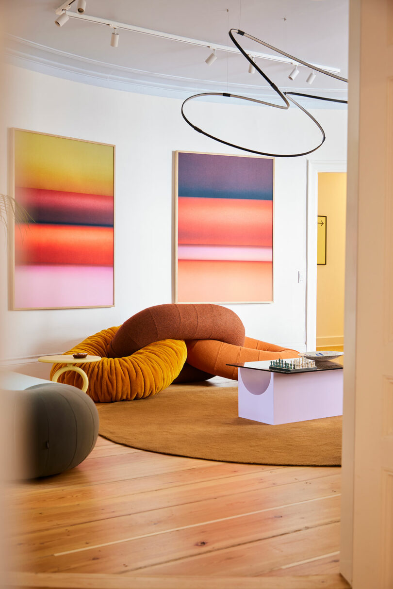 Modern living room with abstract gradient artwork, sculptural orange sofa, low lavender coffee table with chess set, and circular beige rug on wood floor.