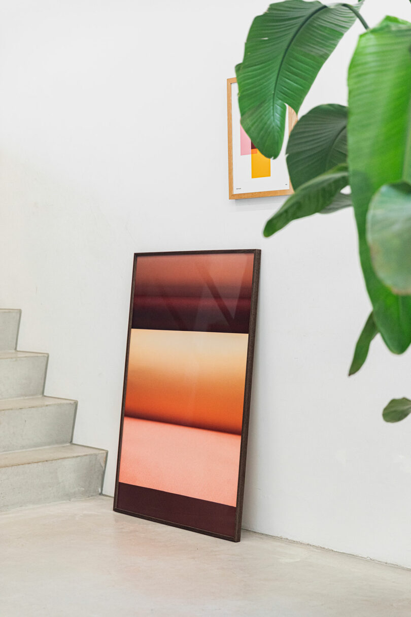 A framed abstract artwork with warm gradient colors leans against a white wall near a staircase, with a large green plant and another framed print visible on the wall above.