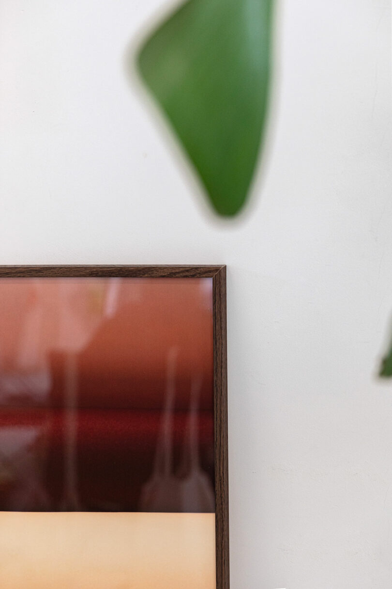 A framed abstract artwork with horizontal bands of brown and beige rests against a white wall; a green leaf is partially visible in the upper right corner.