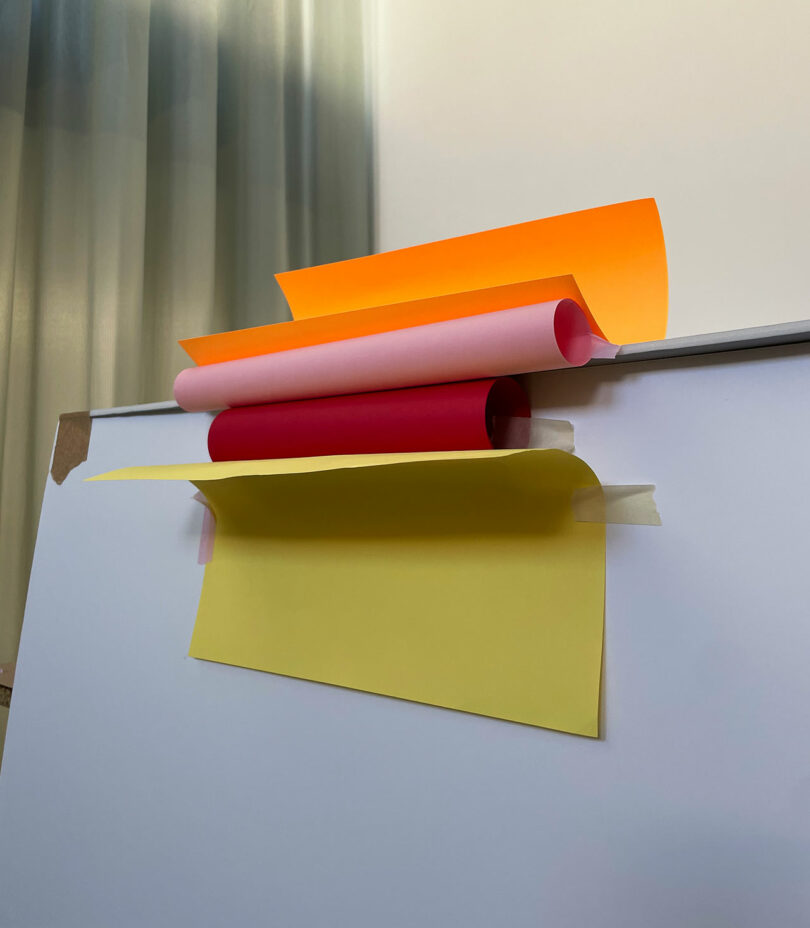 Three sheets of colored paper—orange, red, and yellow—are partially rolled and arranged on top of a whiteboard in a well-lit room.