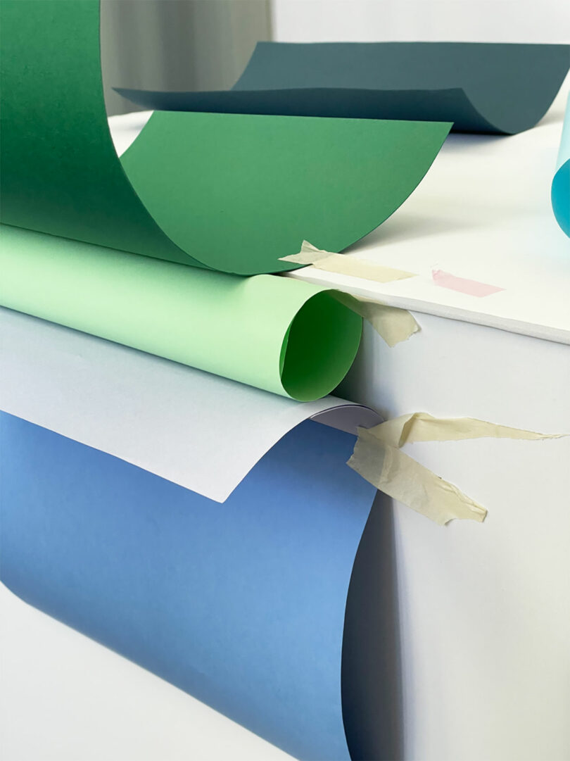 Sheets of green, light green, blue, and white paper are draped and taped to a white surface, with masking tape visible at the edge.