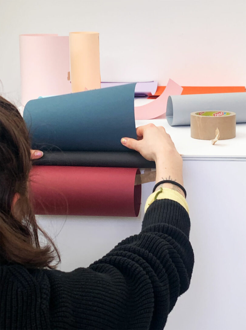 Person arranging sheets of colored paper on a white surface, with rolls of paper and a roll of masking tape in the background.