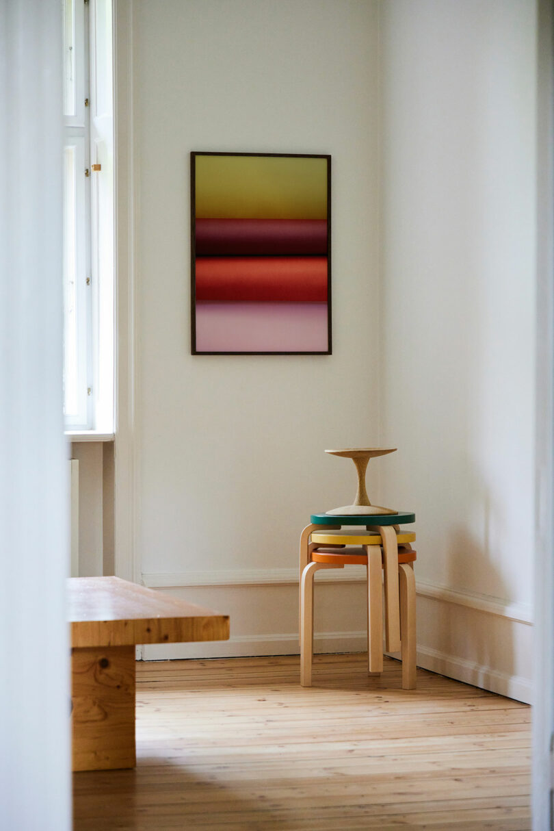 A minimalist room with a wooden bench, a stack of colorful stools, a small tray on top, light wood flooring, and a gradient abstract painting on the wall.