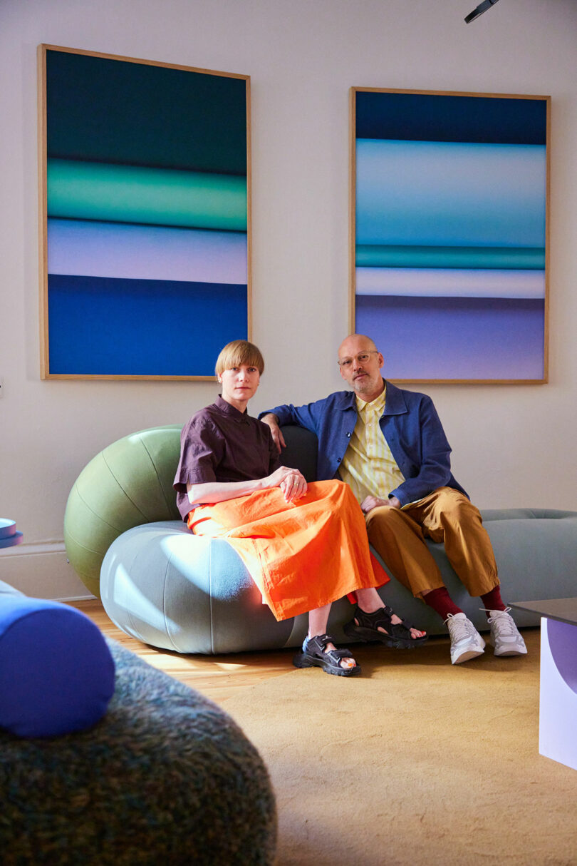 Two people sit on a rounded, modern sofa in a brightly lit room with abstract blue artwork on the wall behind them.