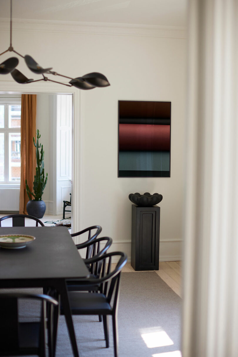 A modern dining room with a black dining table and chairs, abstract wall art, a tall plant in a pot, and a sculptural black vase on a pedestal.