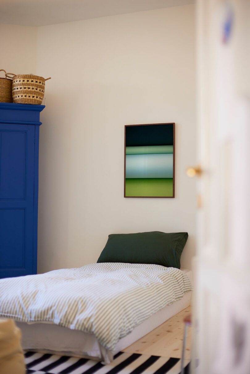 A neatly made single bed with green pillow and striped bedding sits next to a blue wardrobe; abstract artwork hangs above, and woven baskets are on top of the wardrobe.