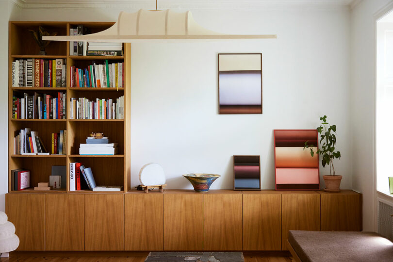 A living room with wooden built-in shelves and cabinets, books, decorative objects, abstract art on the wall, and a potted plant on the right side.