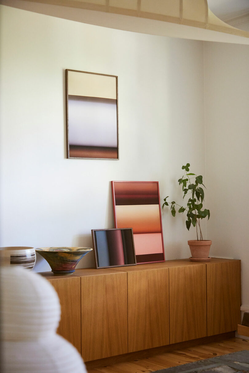 A wooden sideboard with gradient artwork, a potted plant, decorative bowls, and a small framed print against a white wall.