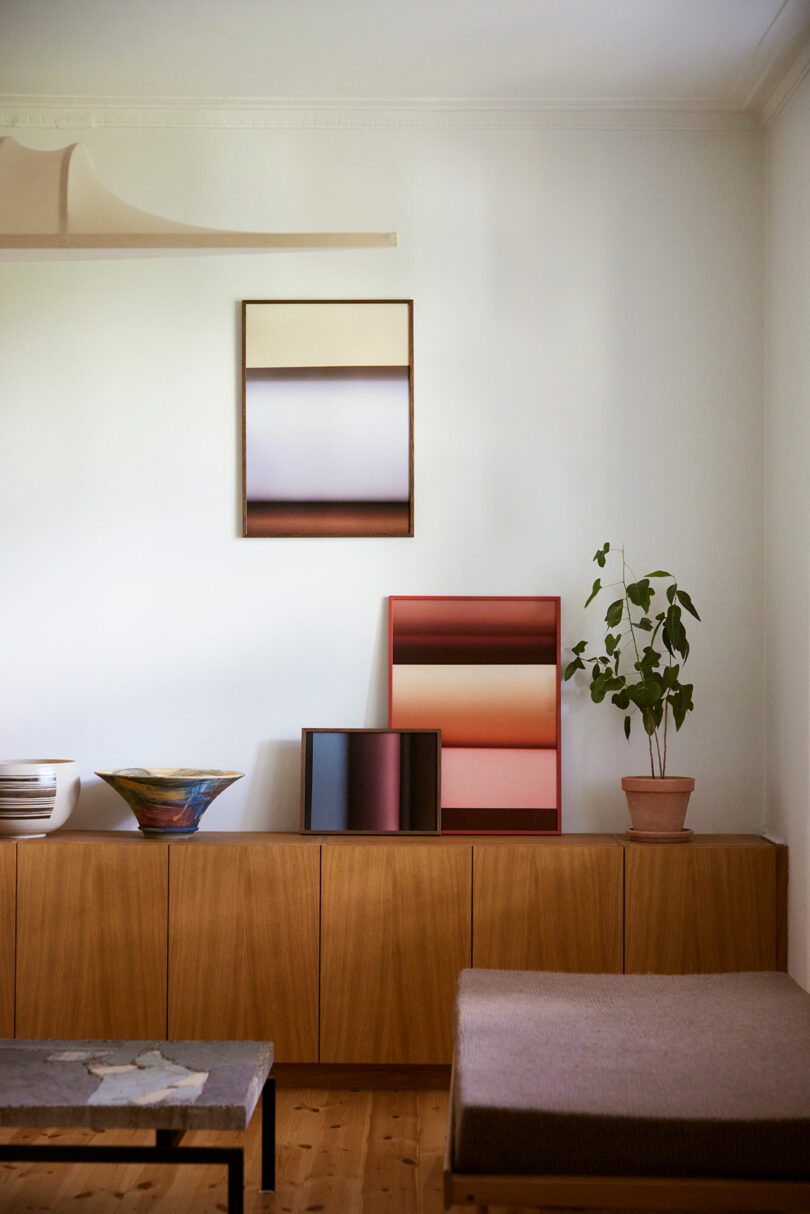 Minimalist living room with a wooden sideboard, abstract gradient art pieces, a potted plant, ceramic bowls, and neutral-toned furniture against a white wall.