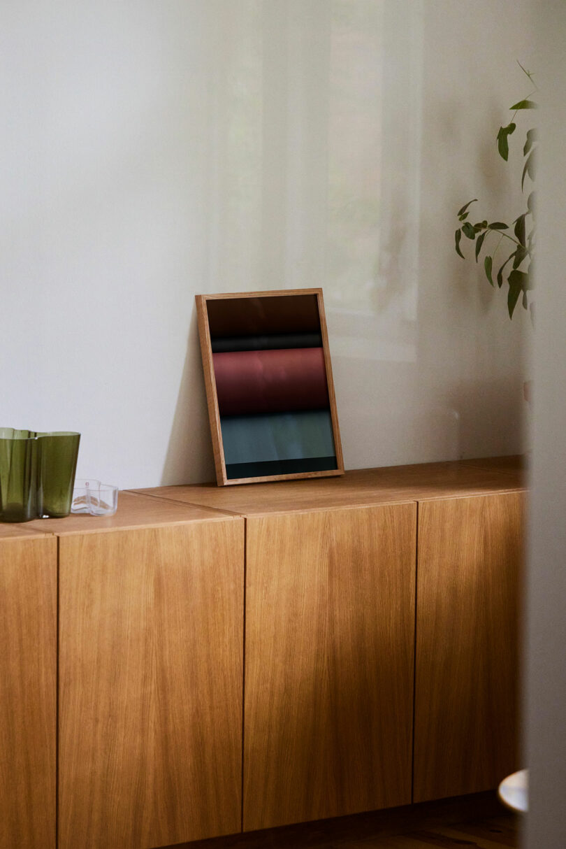 A wooden sideboard with a framed abstract print, two green glasses, and a leafy plant partially visible on the right.