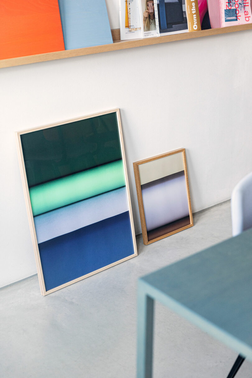 Two abstract art prints in wooden frames lean against a white wall near a light blue table, with books and magazines displayed on a shelf above.