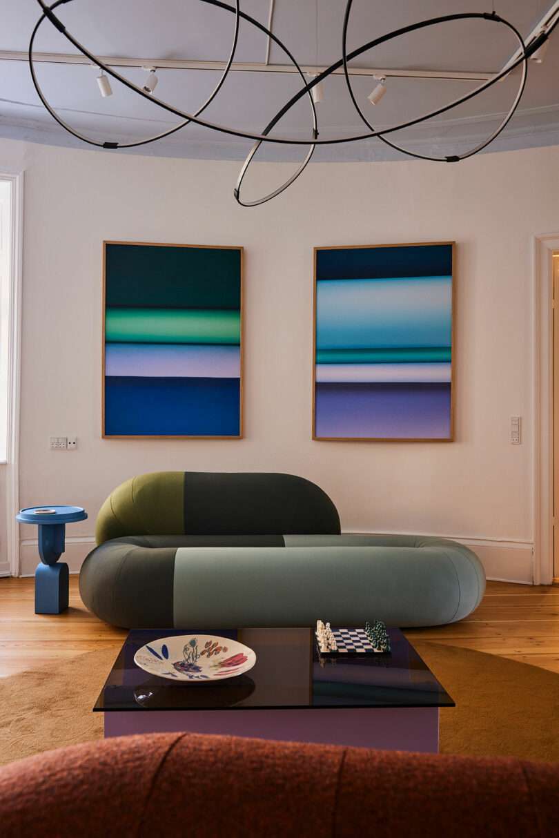 A modern living room with a curved, two-tone sofa, abstract art on the wall, a low coffee table with a chess set and plate, and a sculptural ceiling light fixture.