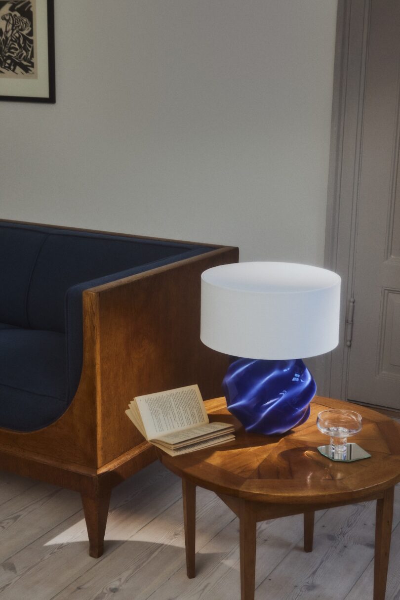 A wooden side table with an open book, a clear ashtray, and a blue sculptural lamp beside a wooden-framed navy sofa in a minimalist room