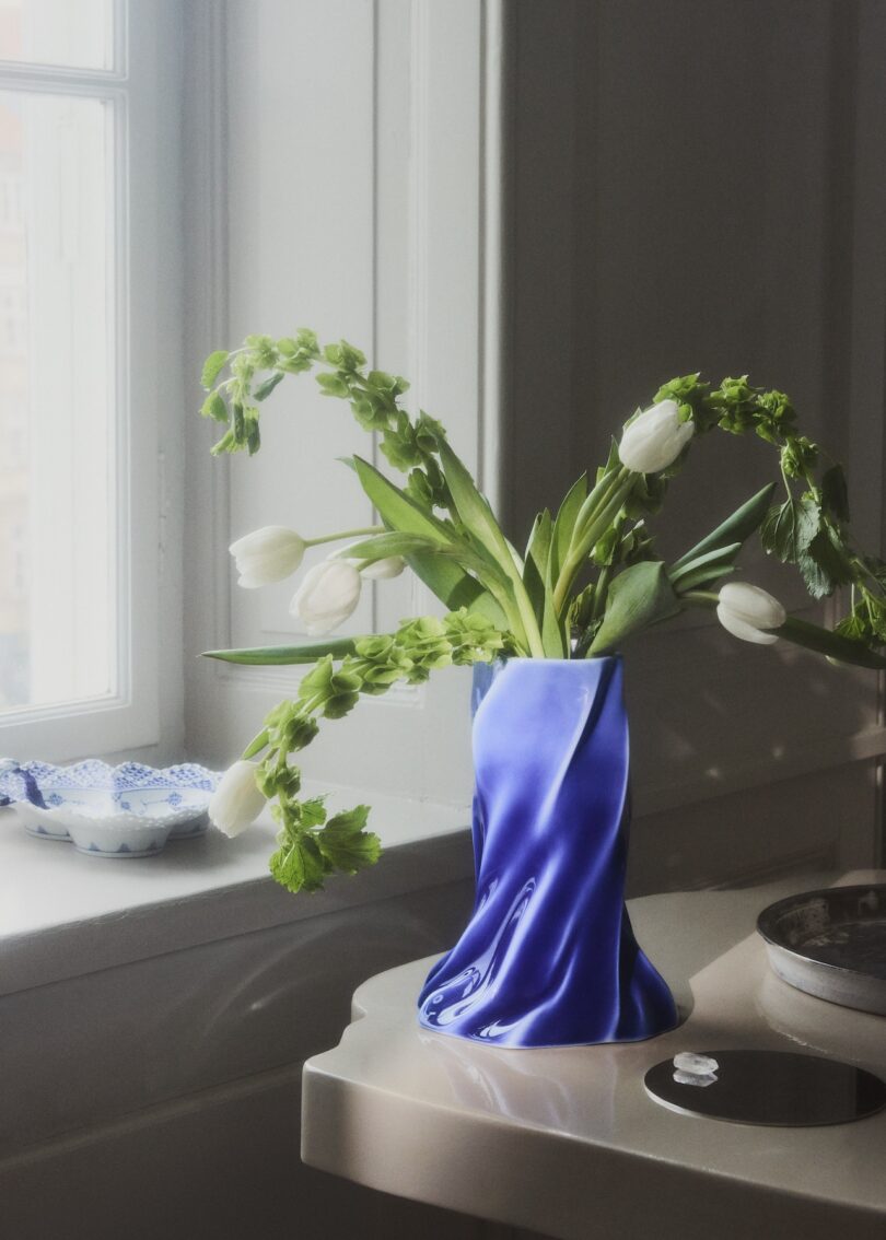 A vase wrapped in blue satin holds white tulips and greenery on a small table by a window with soft natural light