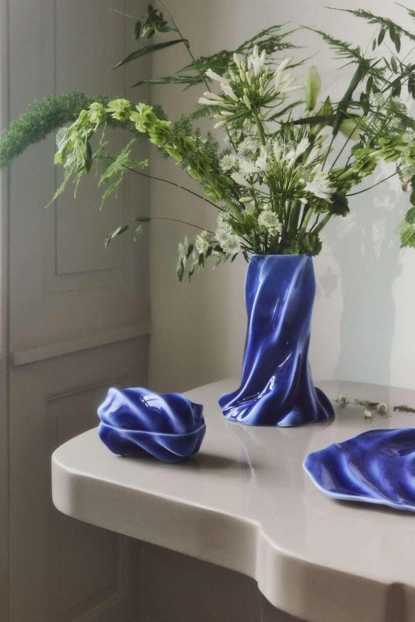 A blue, wavy ceramic vase with green and white flowers sits on a light-colored table next to matching blue ceramic dishes in a bright, minimal room