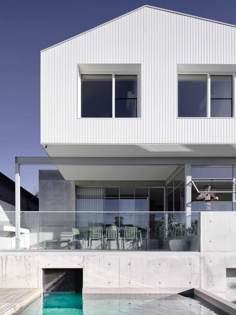 Modern two-story white house with large windows, a covered patio with green chairs, glass railing, and a swimming pool in the foreground under a clear sky.