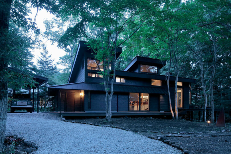 Modern dark-wood house with large windows and warm interior lighting, surrounded by trees and gravel driveway in a wooded setting.