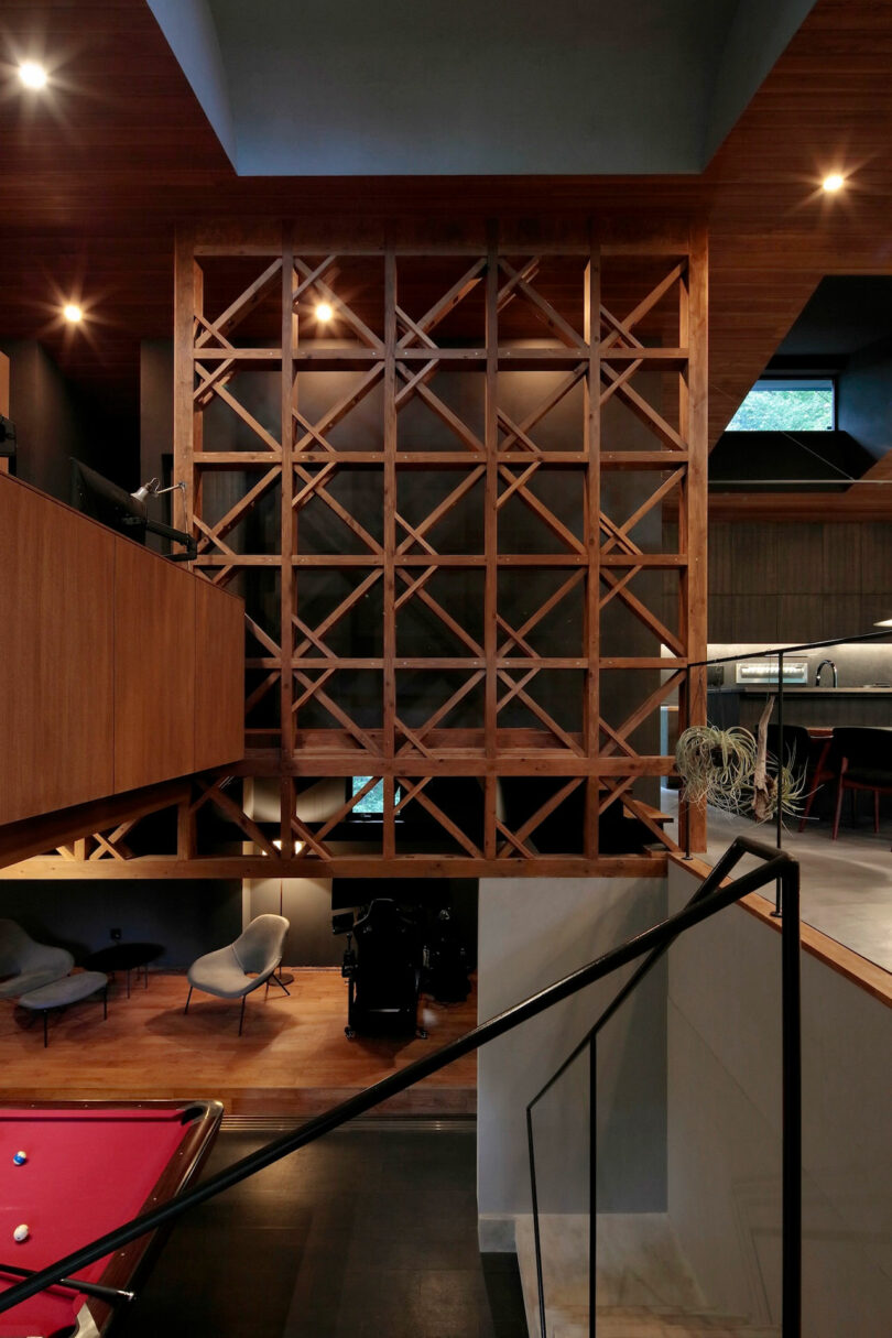 Modern interior featuring a geometric wooden partition, a billiards table, lounge chairs, and an open kitchen with contemporary design elements and warm lighting.