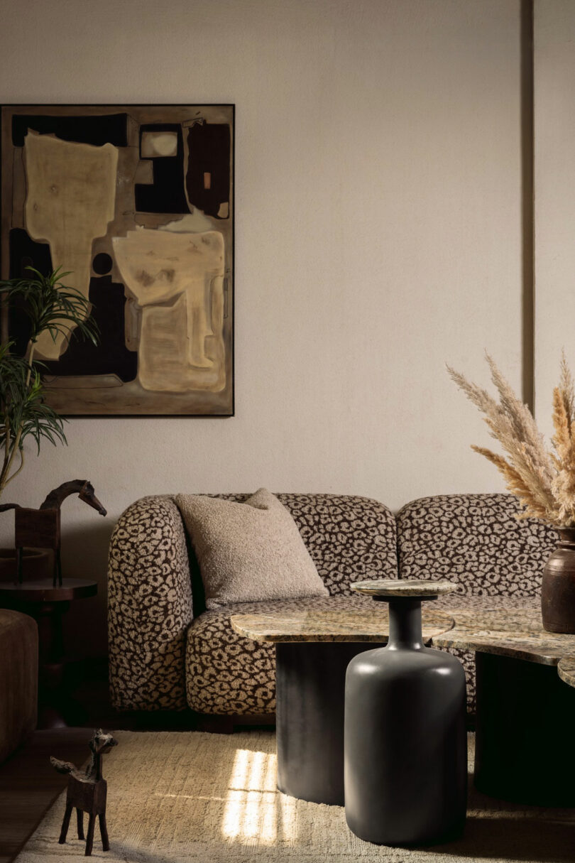 A patterned sofa with a beige pillow sits beside black tables and a vase with pampas grass; abstract art hangs on the wall in a softly lit living room.