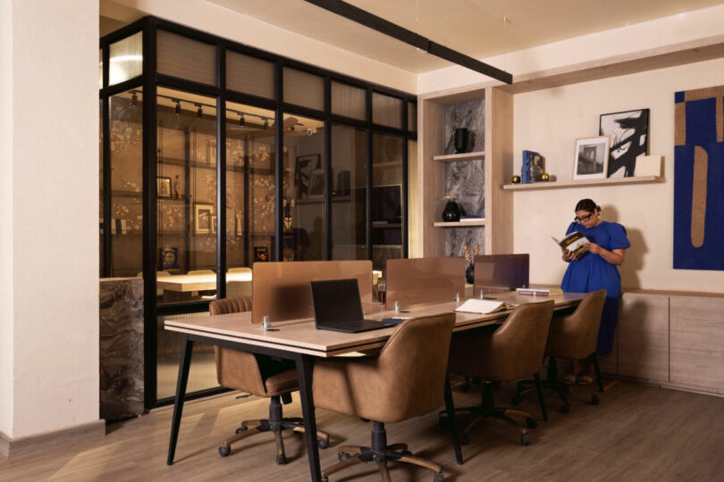 Modern office space with brown chairs and shared desks, laptops on tables, glass-walled meeting room, and a woman in blue standing and reading a book near shelves.