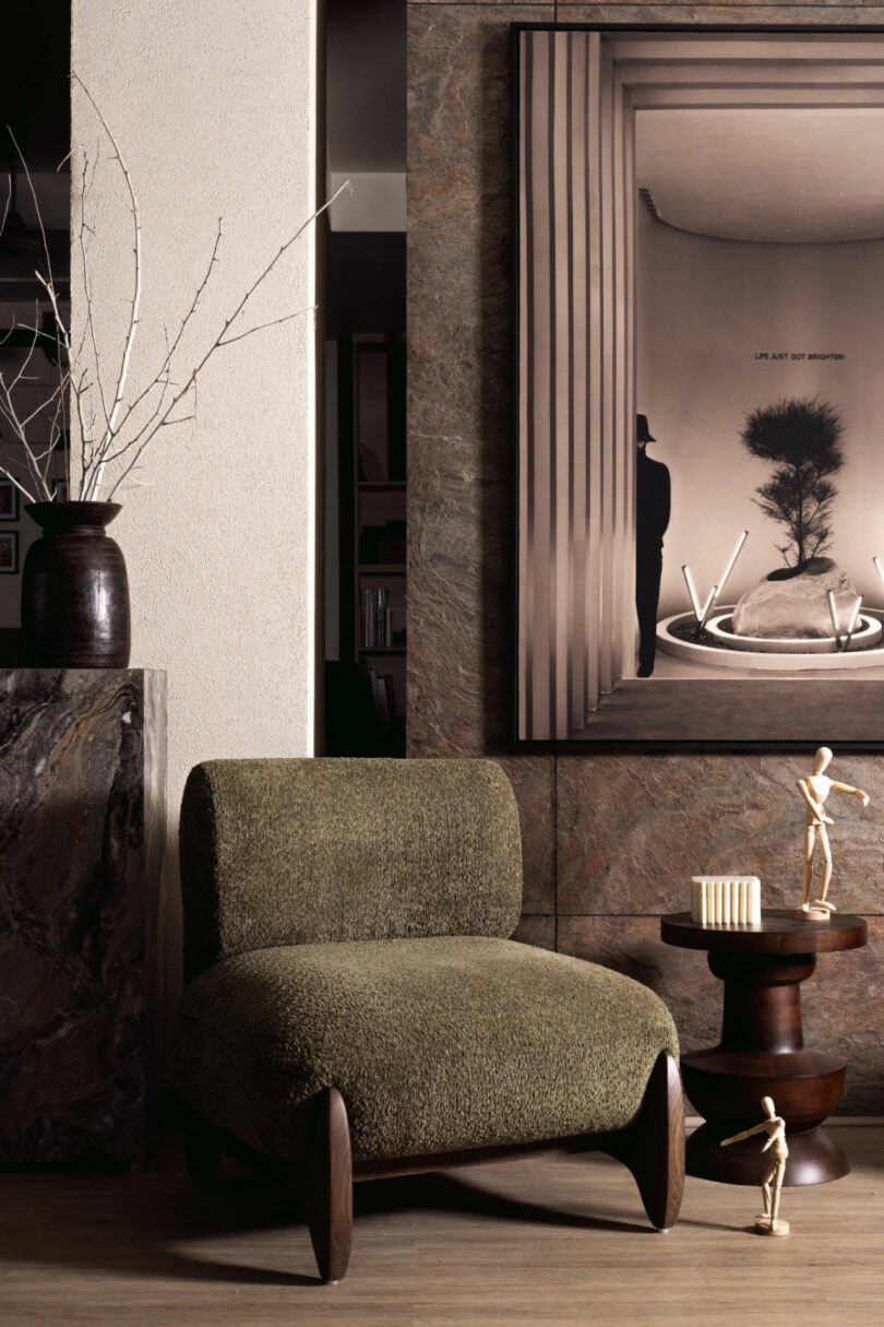 An olive green armchair sits next to a small wooden side table with a candle and wooden figures, beneath framed modern art and a vase with white branches.