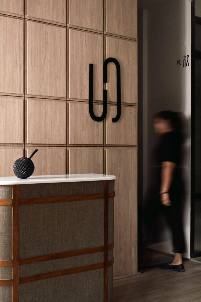 A person in black walks past a tiled wood wall with a modern black wall sculpture near a reception desk and a restroom sign above.