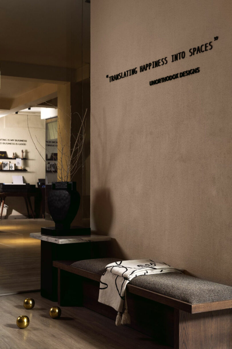 Modern interior with a padded bench, a decorative vase, a minimal patterned throw, and gold spheres on the floor; quote and company name are displayed on the wall.