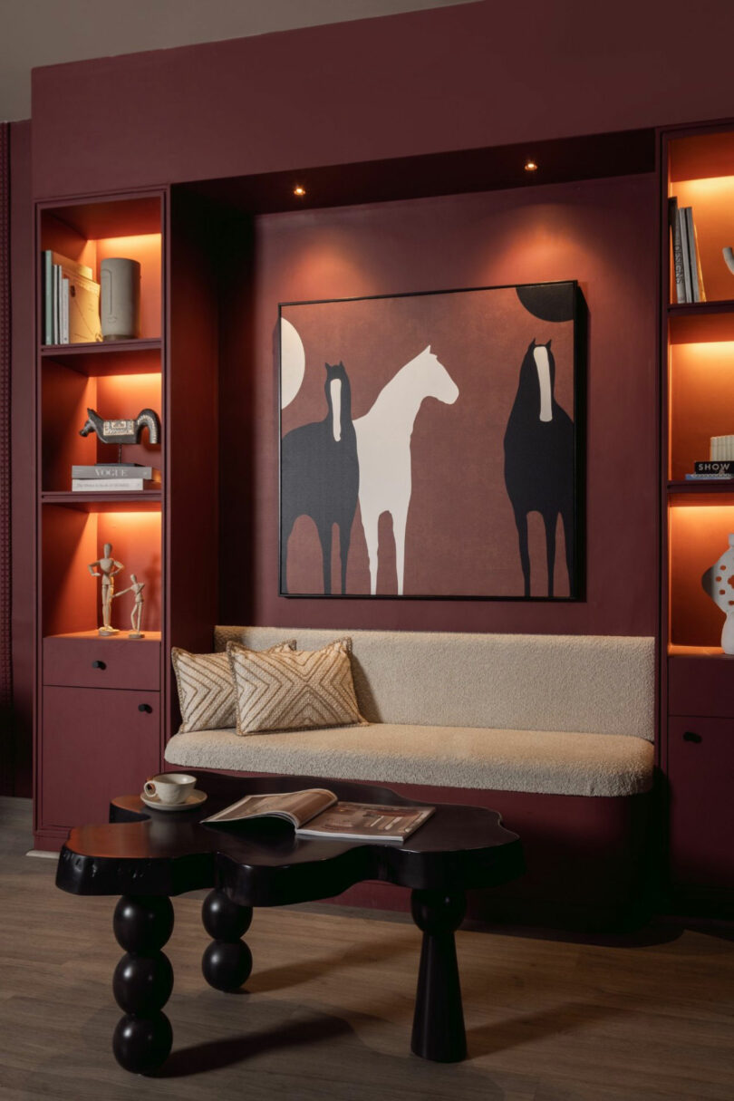 Modern living room with a burgundy accent wall, built-in shelves, beige cushioned bench, abstract horse artwork, and a sculptural black coffee table. Warm ambient lighting throughout.