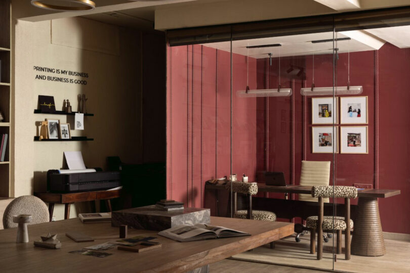 Modern office with a wooden desk in the foreground, a printer and shelves on the left, and a glass-walled meeting room with red walls, framed photos, and two chairs.