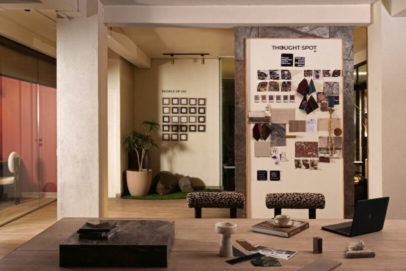 A modern office space with a wooden table, laptop, and materials in the foreground, and inspiration boards, benches, and framed photos on the walls in the background.