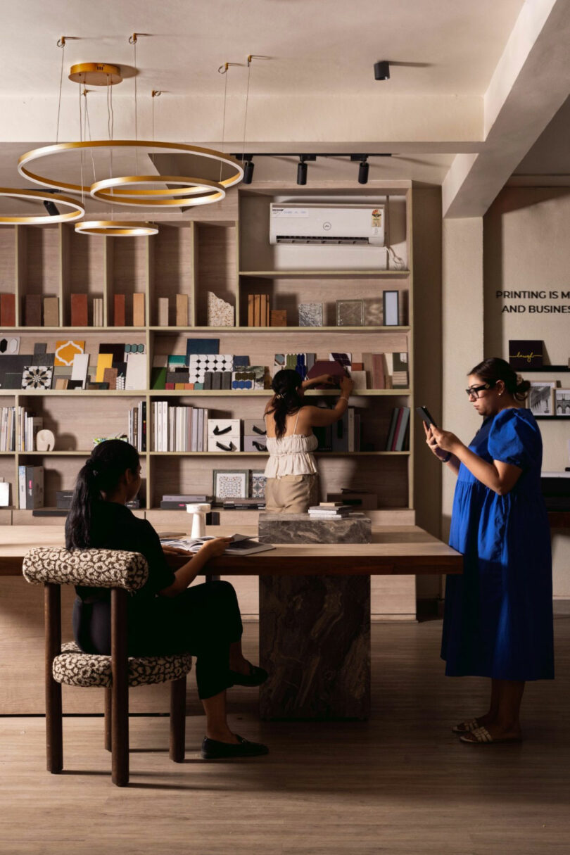 Three women are in a modern library or bookstore; one sits at a table, another arranges books on a shelf, and the third stands taking a photo with her phone.