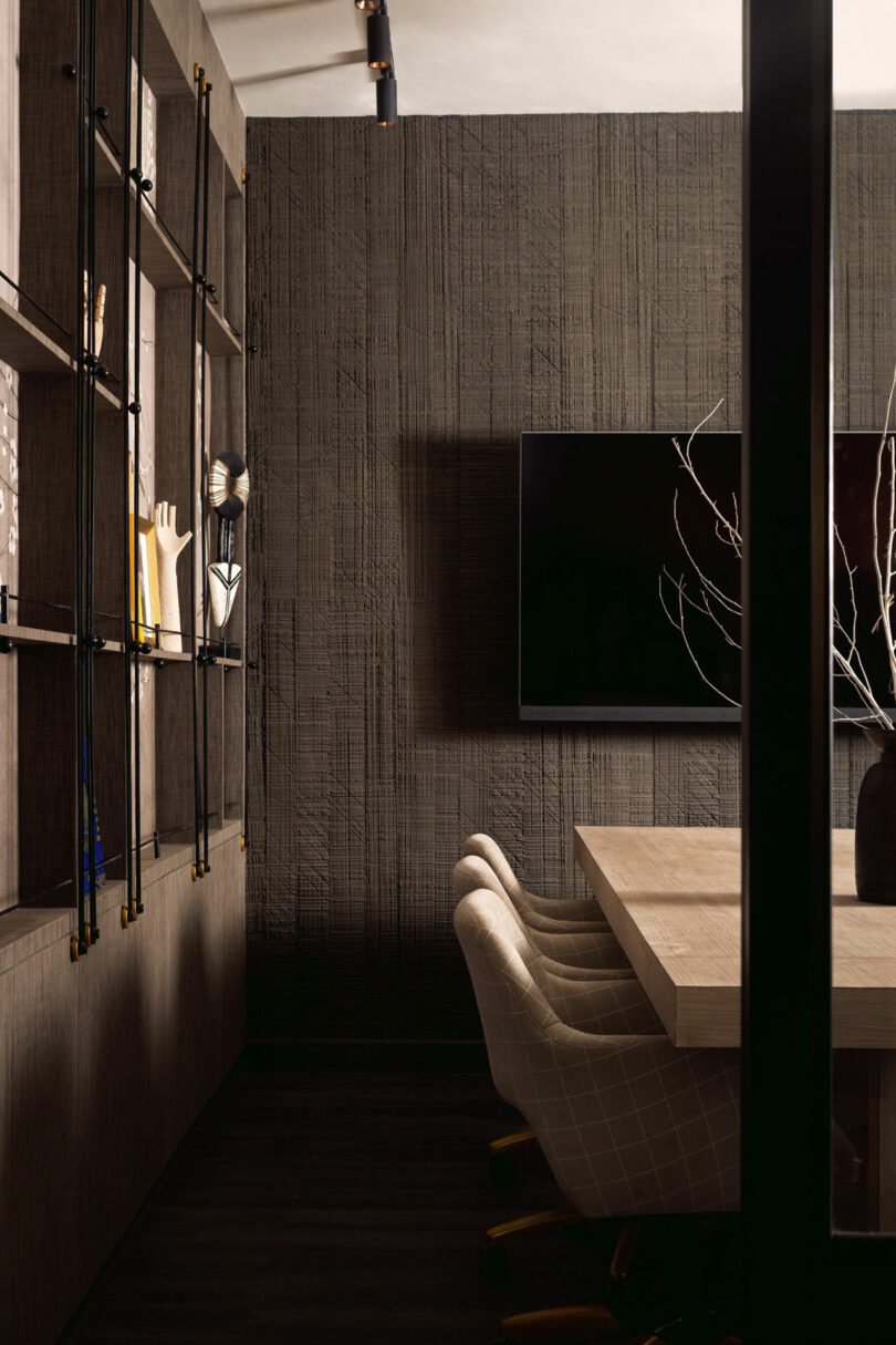 Modern office space with a wood table, four upholstered chairs, wall-mounted TV, and shelving unit displaying decor items against a textured dark wall.