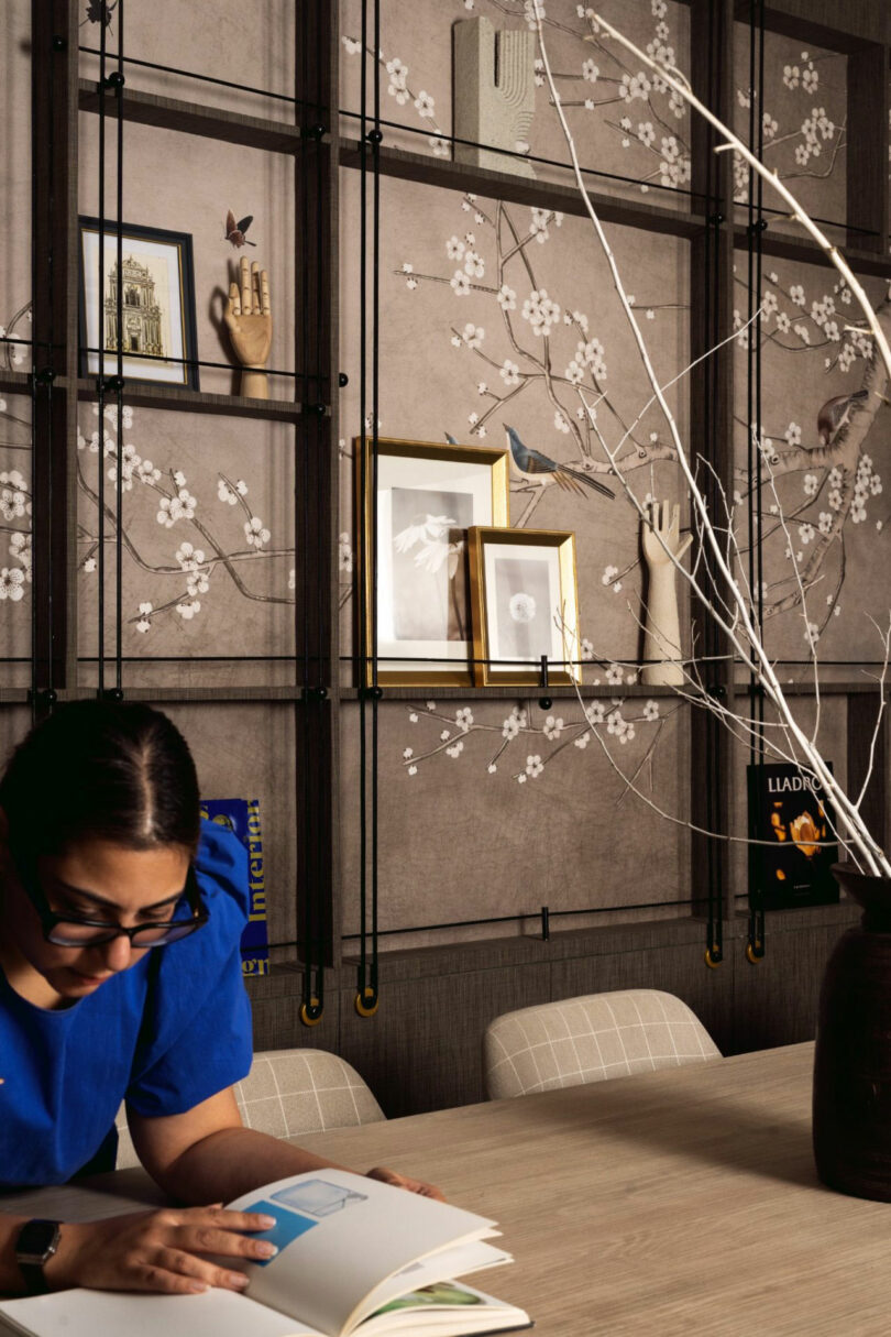 A woman in blue reads at a table in front of a decorative wall with framed photos, art, and floral wallpaper. A vase with bare branches sits on the table.