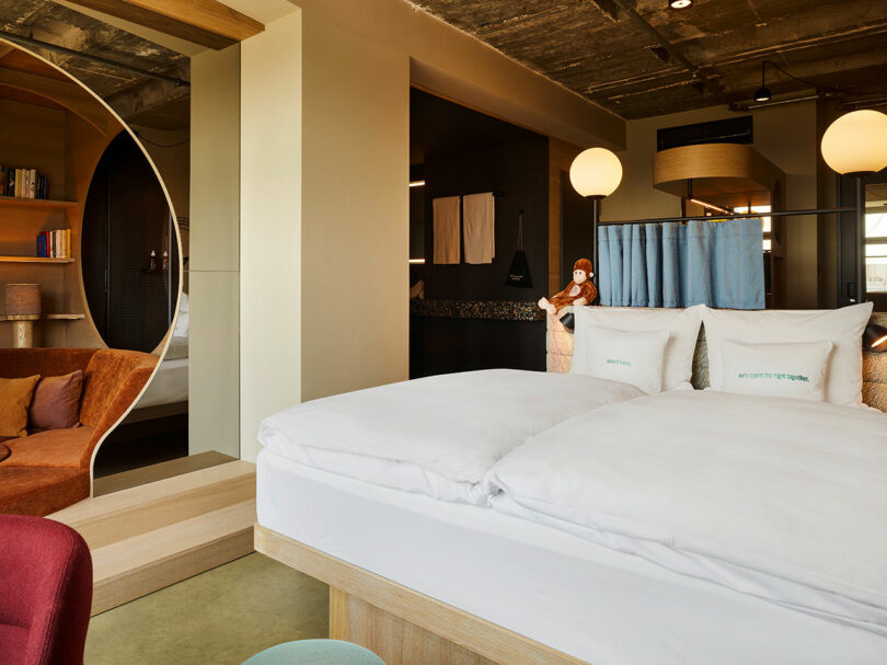 Modern 25hours hotel room with a large bed, white bedding, decorative pillows, a stuffed monkey toy, and a cozy sitting area featuring a bookshelf and round mirror.