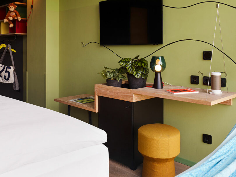 A modern 25hours hotel room with a wooden desk holding a lamp, two potted plants, a notebook, and a cup; a TV on the wall, yellow stool, bed, and hanging blue hammock are visible.
