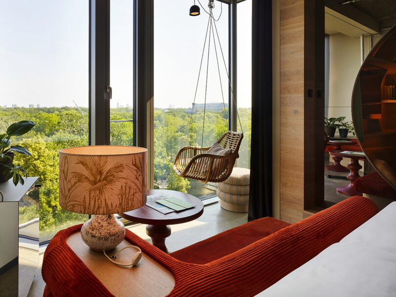 A modern 25hours bedroom with a red couch, a lamp featuring a palm tree design, large windows with views of trees, and a hanging wicker chair by the window.