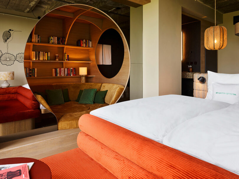 Modern 25hours hotel room with an orange bed, circular wooden partition, built-in bookshelf, brown sofa with green pillows, and abundant natural lighting.