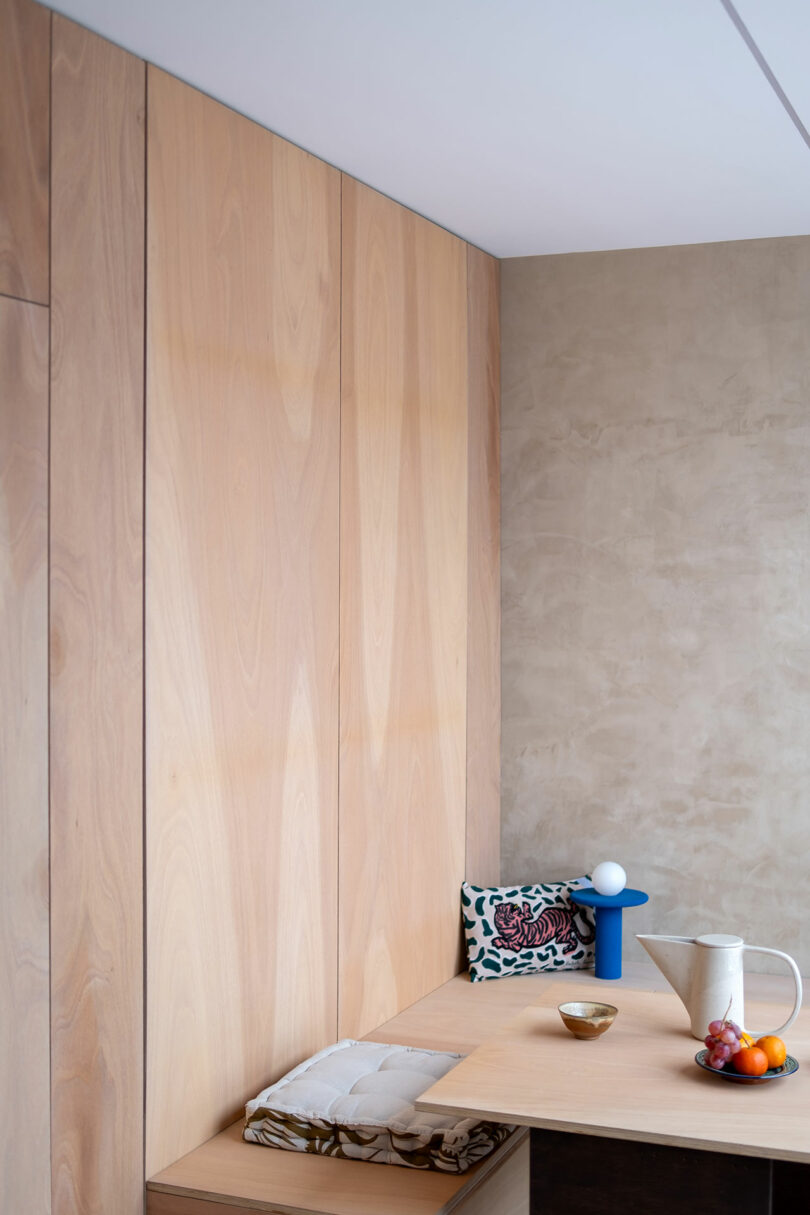 Minimalist dining nook with light wood paneling, a small table with a white pitcher, bowl, fruit, and a blue lamp, plus patterned cushions on bench seating.