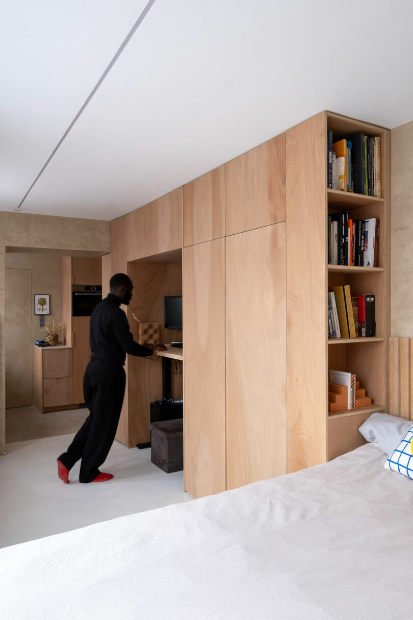 A person stands at a built-in wooden desk with shelves and cabinets, working in a minimalist bedroom with light wood furniture and neutral walls.