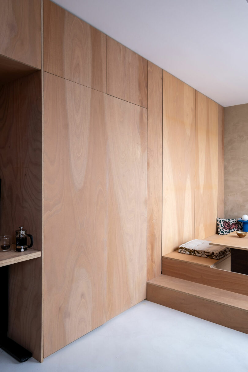 Minimalist interior with light wood panel walls, a built-in wooden bench with cushions, and a small shelf holding a French press and glass on the left side.