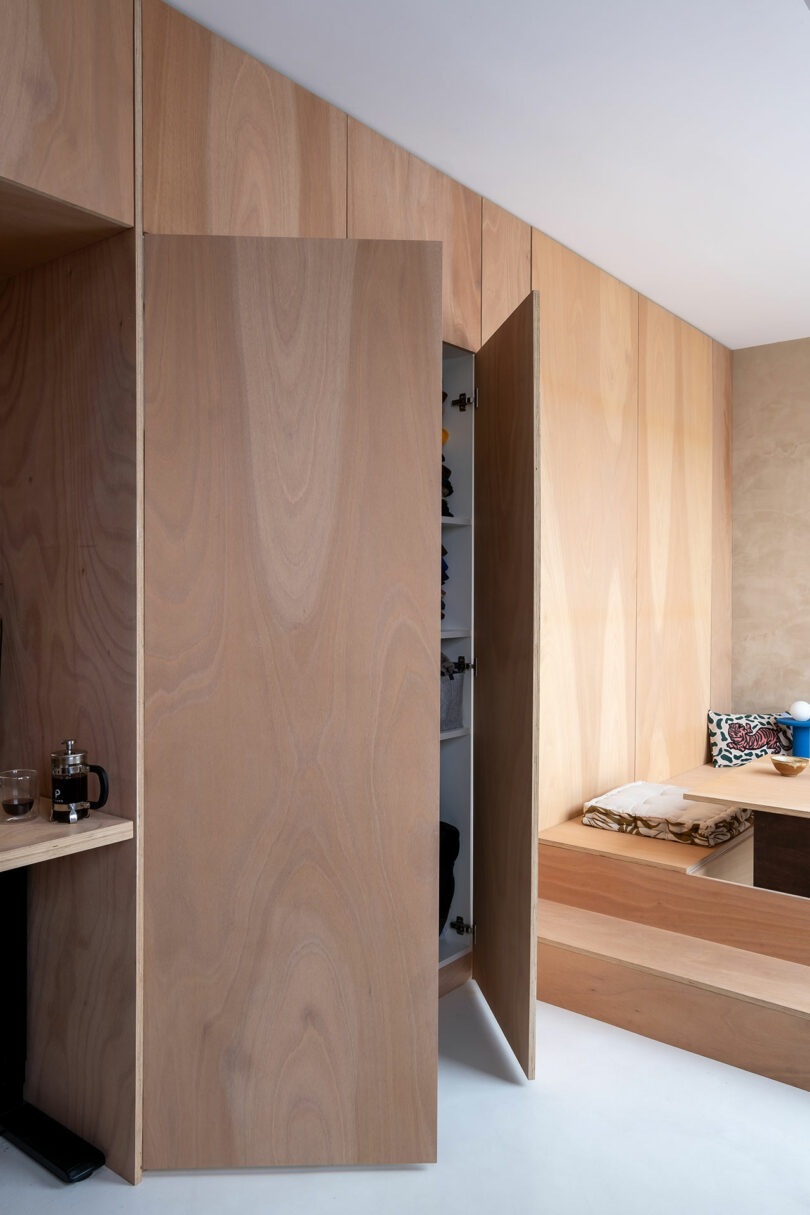 Minimalist wooden closet with an open door reveals shelves with folded clothing; a bed with cushions is visible to the right.