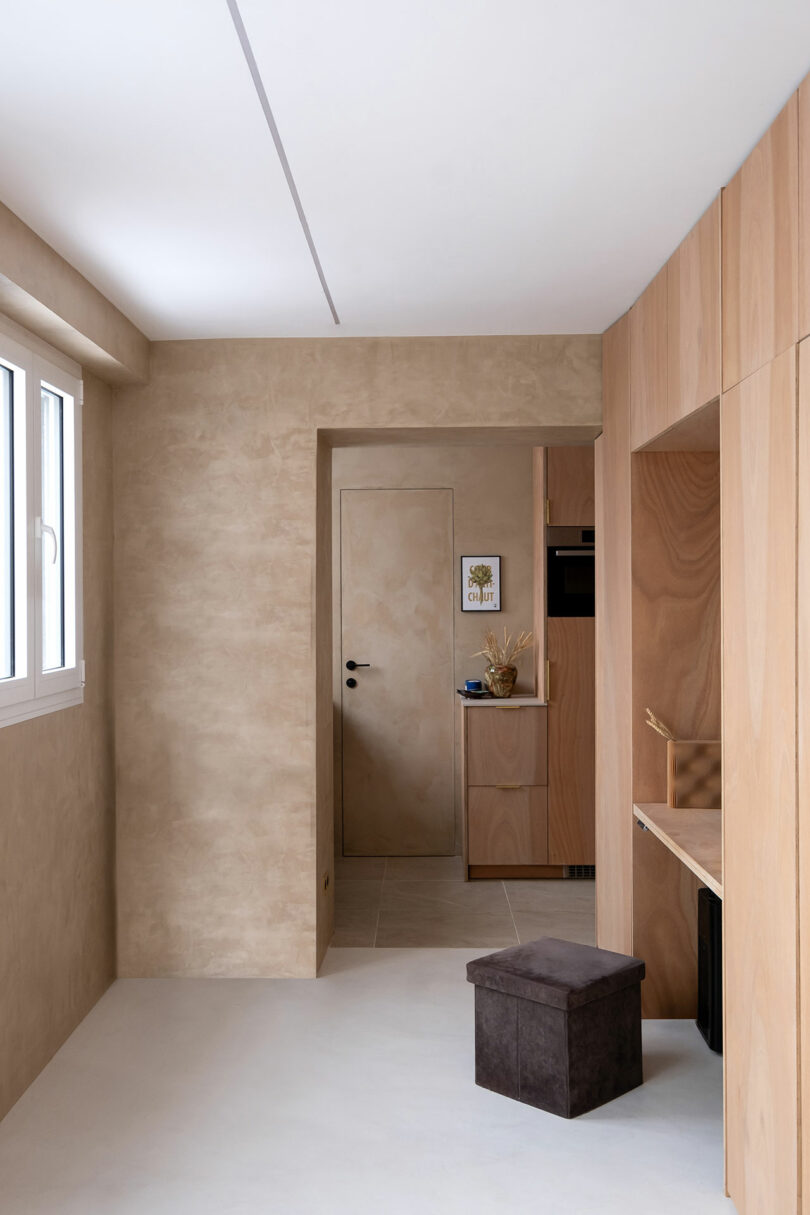 Minimalist room with light beige walls, built-in wood cabinetry, a small dark storage ottoman, and a door leading to another space with framed art on the wall.