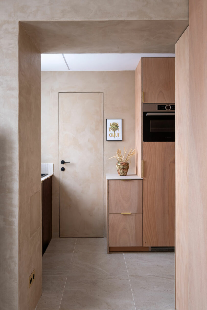 Minimalist kitchen with light wood cabinets, beige walls, a closed door, built-in appliances, and a small framed artwork above a vase on the countertop.