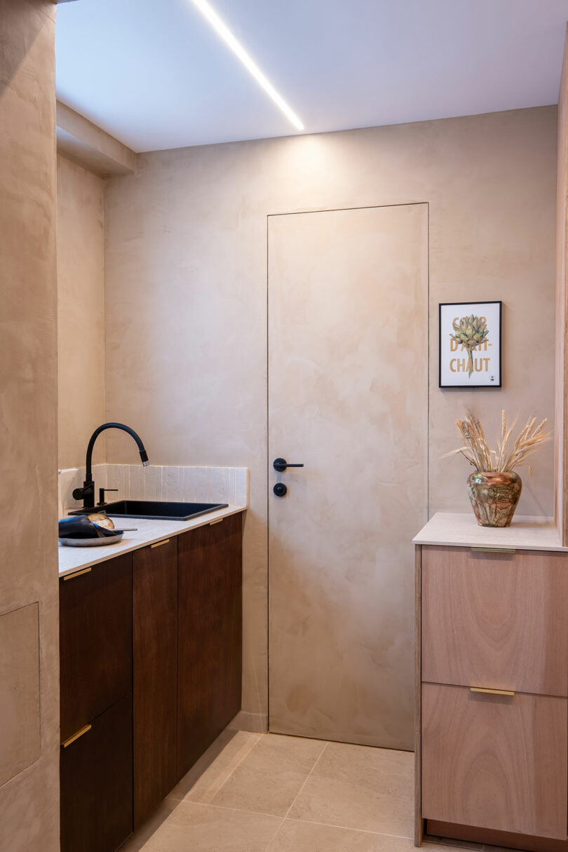 A small modern bathroom with beige walls, a black faucet over a dark wood vanity, a light wood cabinet, and framed wall art above a vase with dried plants.