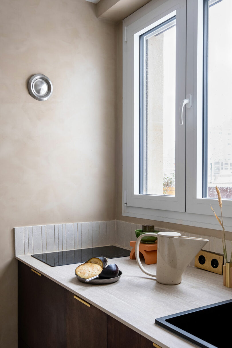 A minimalist kitchen countertop with a cutting board holding sliced bread and an eggplant, a beige pitcher, two small potted plants, and a stovetop beneath a large window.