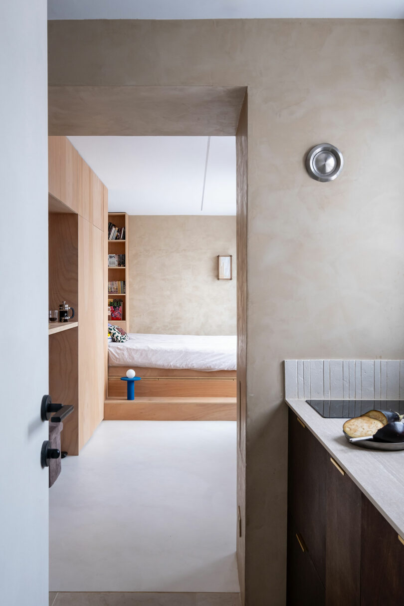 View from a kitchen into a minimalist bedroom with light wood furnishings, built-in shelves, and a neatly made bed.